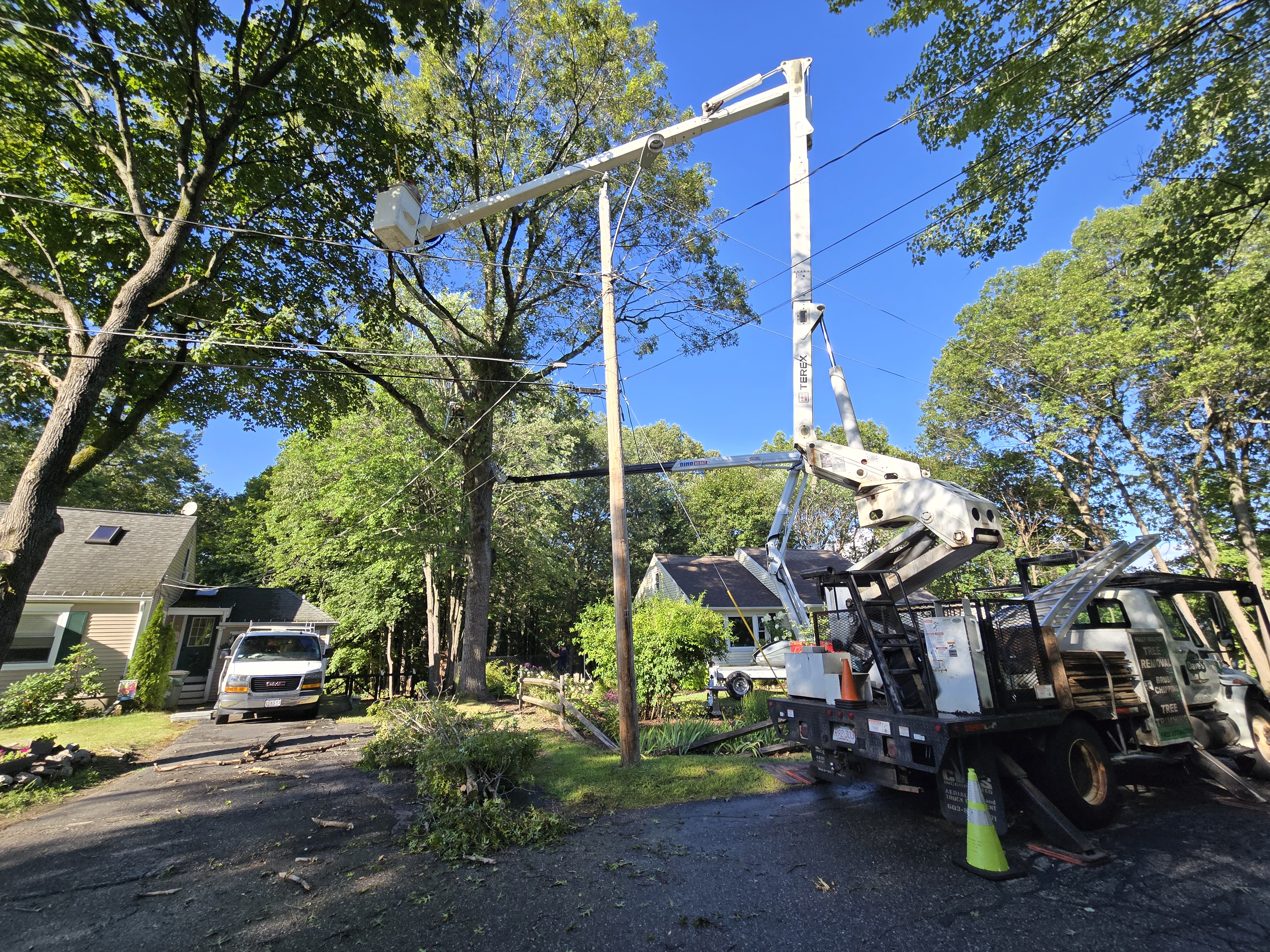White utility service truck with extended aerial lift on a residential street surrounded by mature trees and houses, lift frame visible against the blue sky for tree trimming or similar work.